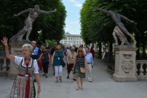 Tourists explore a tree-lined path with statues leading to a grand building.