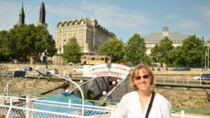 Woman smiling near a boat docked by a bridge in a sunny city park.