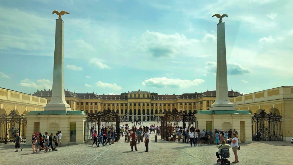 Crowd gathered outside a grand historic palace with classical architecture.