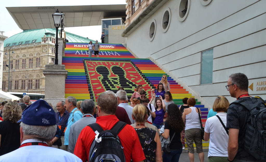 Crowd ascending colorful rainbow stairs with bold black text outdoors.