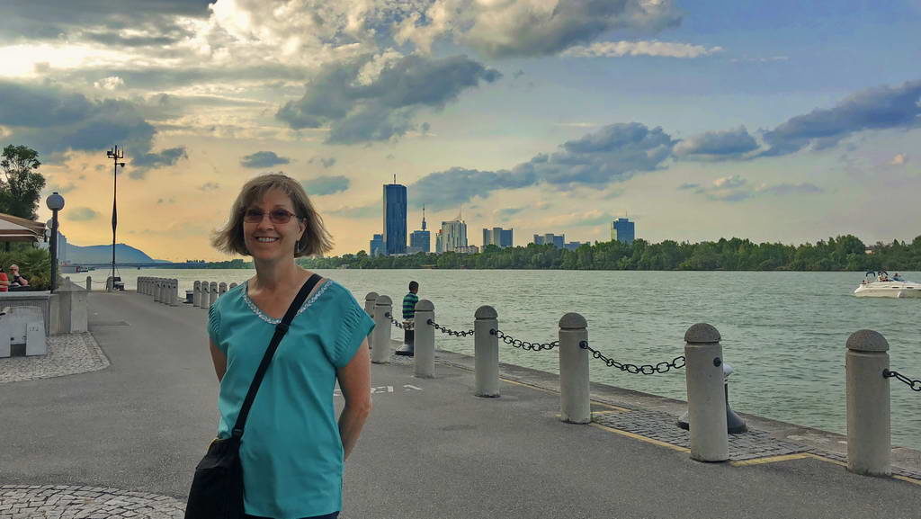 Woman in a turquoise shirt stands by a lakeside with a city skyline behind her.