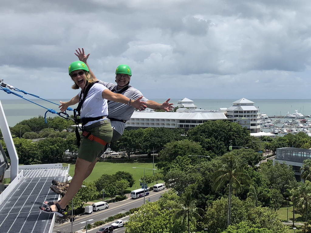 Two people ziplining over a green landscape with cruise ships in the background.