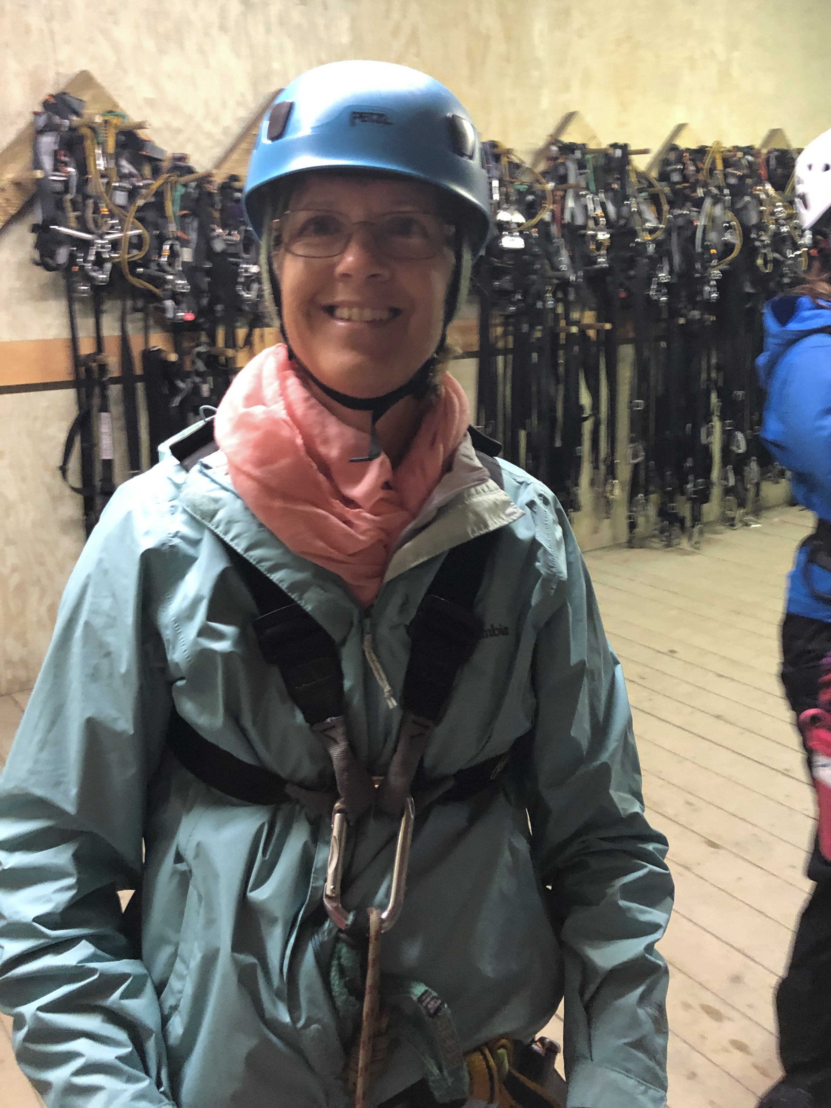 Smiling person in outdoor gear and helmet indoors with equipment in background.
